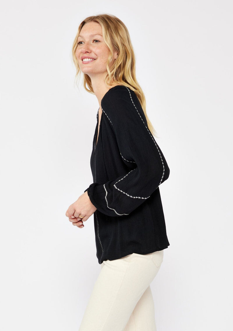 [Color: Black/Natural] A blonde model wearing an embroidered black bohemian blouse. With a split v neckline with ties, long raglan sleeves, and an elastic cuff. A relaxed fit boho top styled with beige denim for the ultimate fall ensemble.