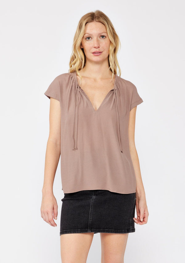 [Color: Mocha] A blonde model wearing a relaxed fit  brown mocha top. With self neck ties, ruched split v neckline, and short dolman sleeves. Styled with a cute black denim skirt for a laid back look. 