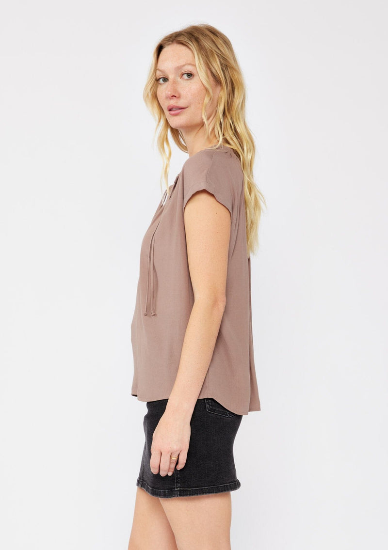 [Color: Mocha] A blonde model wearing a relaxed fit brown mocha top. With self neck ties, ruched split v neckline, and short dolman sleeves. Styled with a cute black denim skirt for a laid back look.