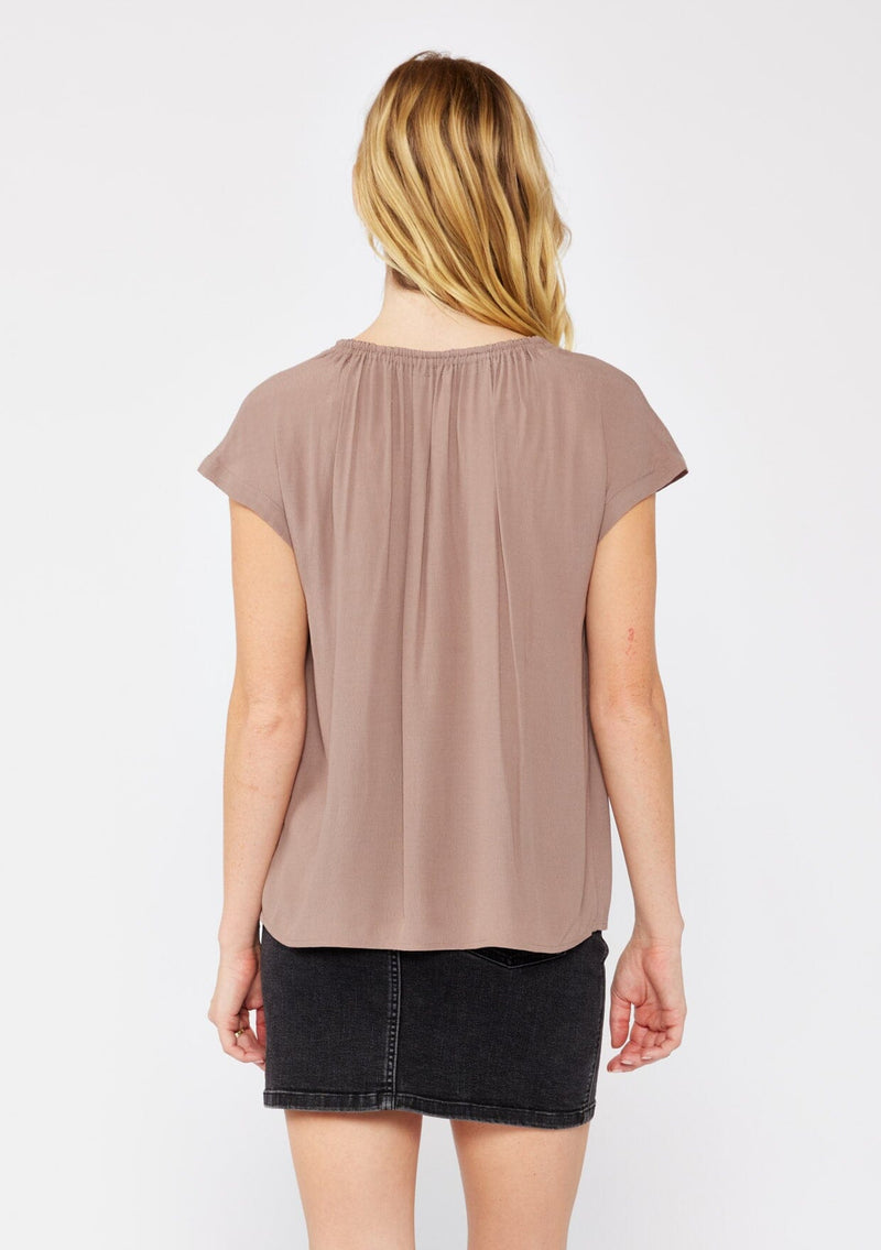 [Color: Mocha] A blonde model wearing a relaxed fit brown mocha top. With self neck ties, ruched split v neckline, and short dolman sleeves. Styled with a cute black denim skirt for a laid back look.