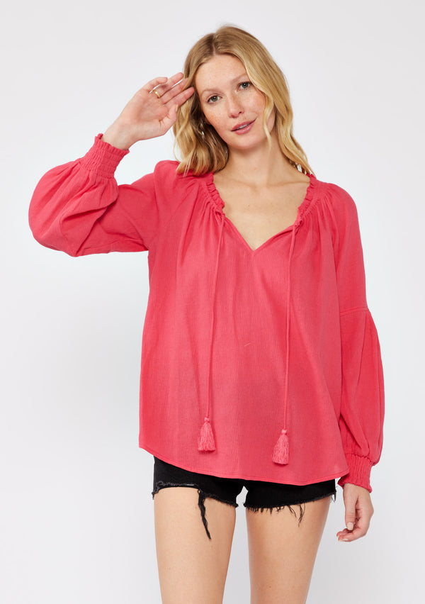 [Color: Watermelon] A blonde model wearing a classic lightweight sheer coral pink peasant top. With a ruffled neckline, tassel ties, long voluminous bishop sleeves, and a relaxed flowy fit.