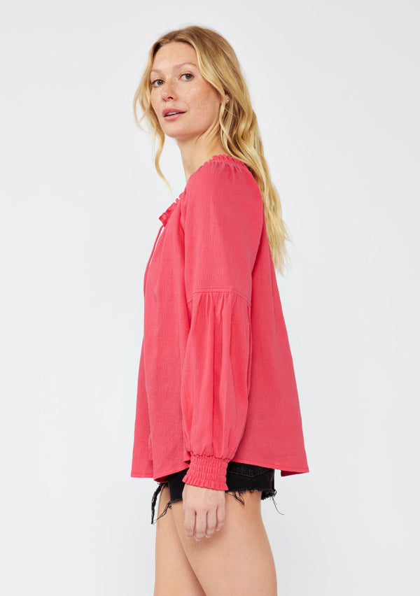[Color: Watermelon] A blonde model wearing a classic lightweight sheer coral pink peasant top. With a ruffled neckline, tassel ties, long voluminous bishop sleeves, and a relaxed flowy fit.