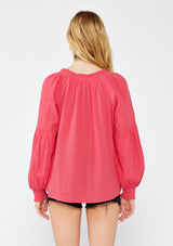 [Color: Watermelon] A blonde model wearing a classic lightweight sheer coral pink peasant top. With a ruffled neckline, tassel ties, long voluminous bishop sleeves, and a relaxed flowy fit.