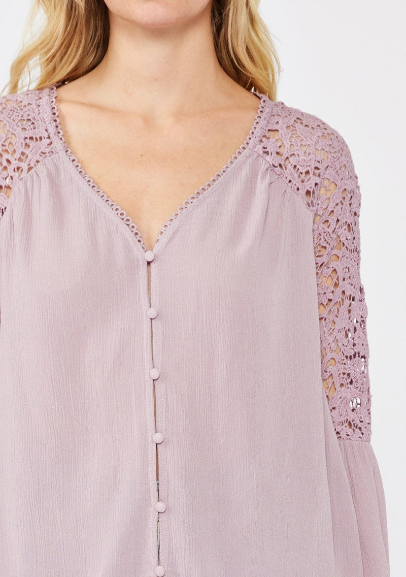 [Color: Dusty Lilac] An image of a blonde model wearing a light purple bohemian blouse with a v neckline with lace design, a self covered button front, and three quarter length bell sleeves with sheer crochet lace details.