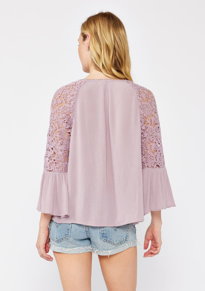 [Color: Dusty Lilac] An image of a blonde model wearing a light purple bohemian blouse with a v neckline with lace design, a self covered button front, and three quarter length bell sleeves with sheer crochet lace details.
