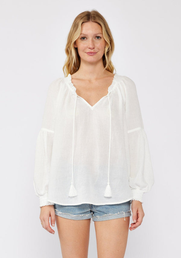 [Color: White] A blonde model wearing a classic lightweight sheer white peasant top. With a ruffled neckline, tassel ties, long voluminous bishop sleeves, and a relaxed flowy fit. 