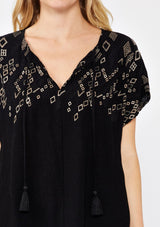 [Color: Black/Cement] A front detail image pf a blonde model standing outside wearing an black boxy fit bohemian fall top with brown embroidered detail. With short dolman sleeves, a split v neckline, and tassel ties.