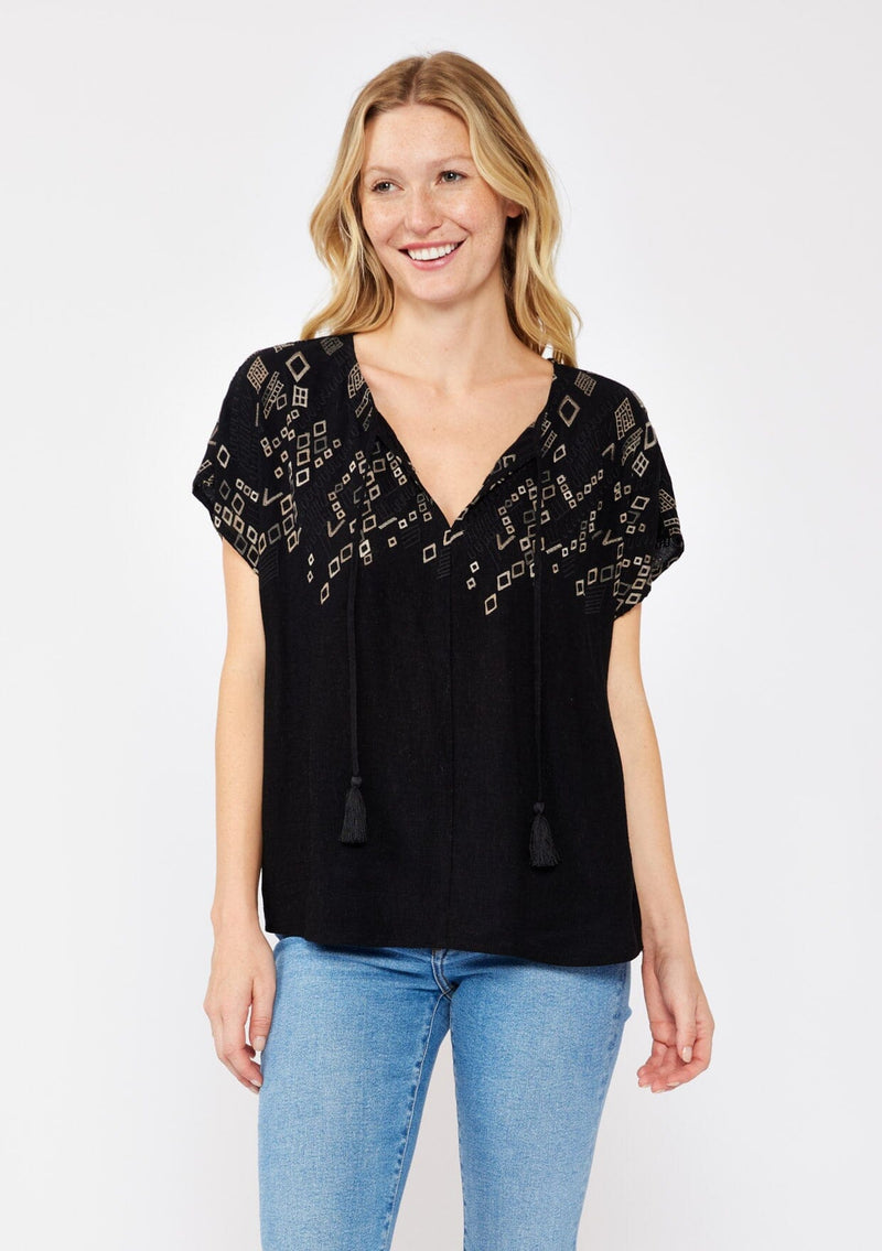 [Color: Black/Cement] A front facing image pf a blonde model standing outside wearing an black boxy fit bohemian fall top with brown embroidered detail. With short dolman sleeves, a split v neckline, and tassel ties. 