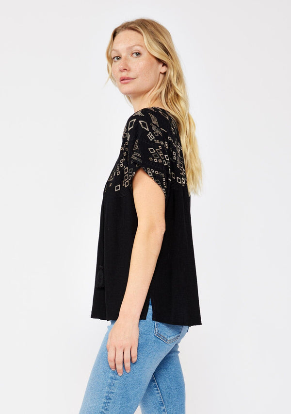 [Color: Black/Cement] A side facing image pf a blonde model standing outside wearing an black boxy fit bohemian fall top with brown embroidered detail. With short dolman sleeves, a split v neckline, and tassel ties.
