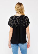 [Color: Black/Cement] A back facing image pf a blonde model standing outside wearing an black boxy fit bohemian fall top with brown embroidered detail. With short dolman sleeves, a split v neckline, and tassel ties.