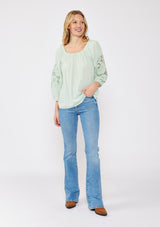 [Color: Dusty Sage] A brunette model wearing a casual green minty blouse with paisley lace detail. A summer top with an elastic neckline for on and off shoulder styling and three quarter length sleeves with elastic cuffs. A relaxed a flowy style perfect for casual outings.