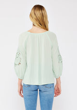 [Color: Dusty Sage] A brunette model wearing a casual green minty blouse with paisley lace detail. A summer top with an elastic neckline for on and off shoulder styling and three quarter length sleeves with elastic cuffs. A relaxed a flowy style perfect for casual outings.