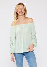 [Color: Dusty Sage] A brunette model wearing a casual green minty blouse with paisley lace detail. A summer top with an elastic neckline for on and off shoulder styling and three quarter length sleeves with elastic cuffs. A relaxed a flowy style perfect for casual outings.