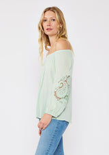 [Color: Dusty Sage] A brunette model wearing a casual green minty blouse with paisley lace detail. A summer top with an elastic neckline for on and off shoulder styling and three quarter length sleeves with elastic cuffs. A relaxed a flowy style perfect for casual outings.