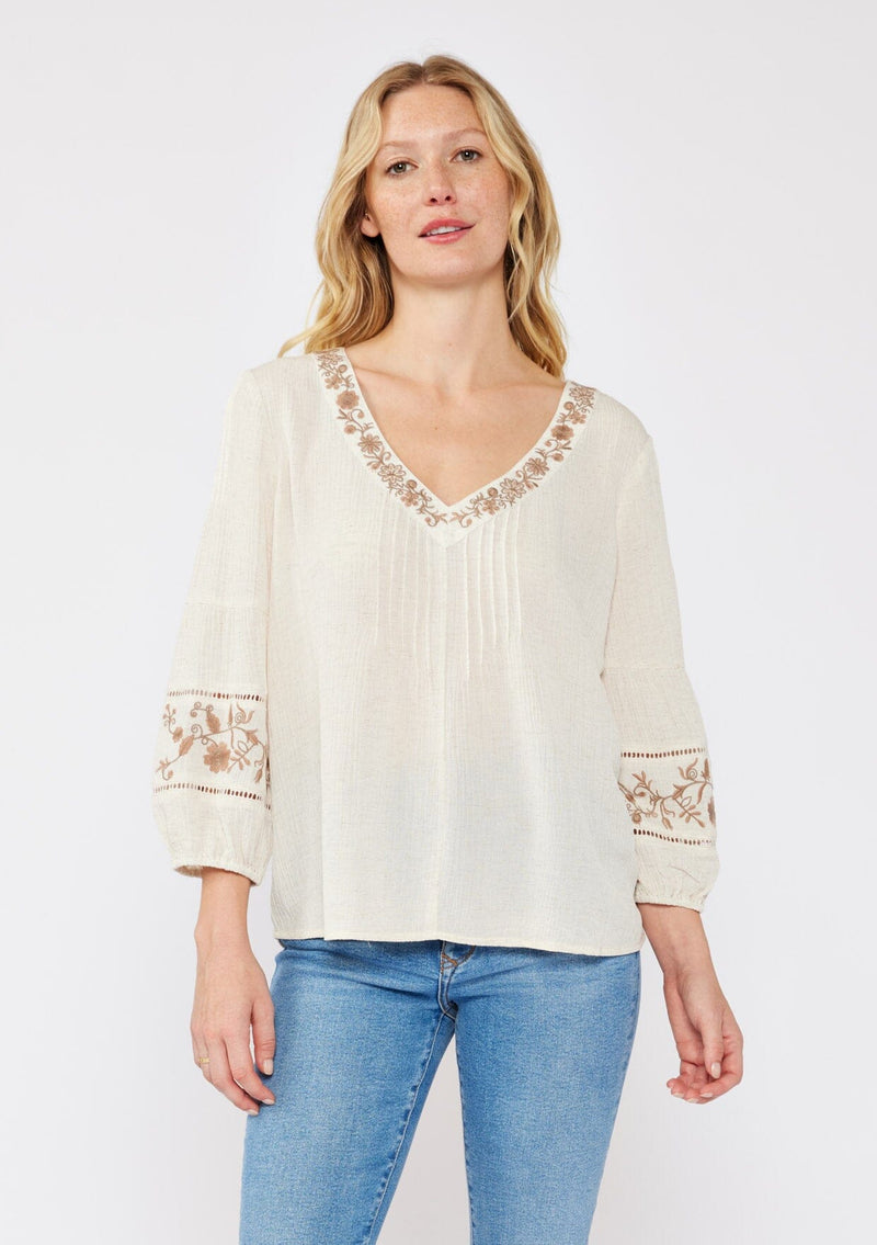 [Color: Natural/Brown] An image of a blonde model wearing an ultra bohemian blouse with embroidered details. A beige natural tone blouse with a v neckline, floral embroidery, and long sleeves with elastic cuffs. A relaxed fit fall season top paired with denim jeans. 