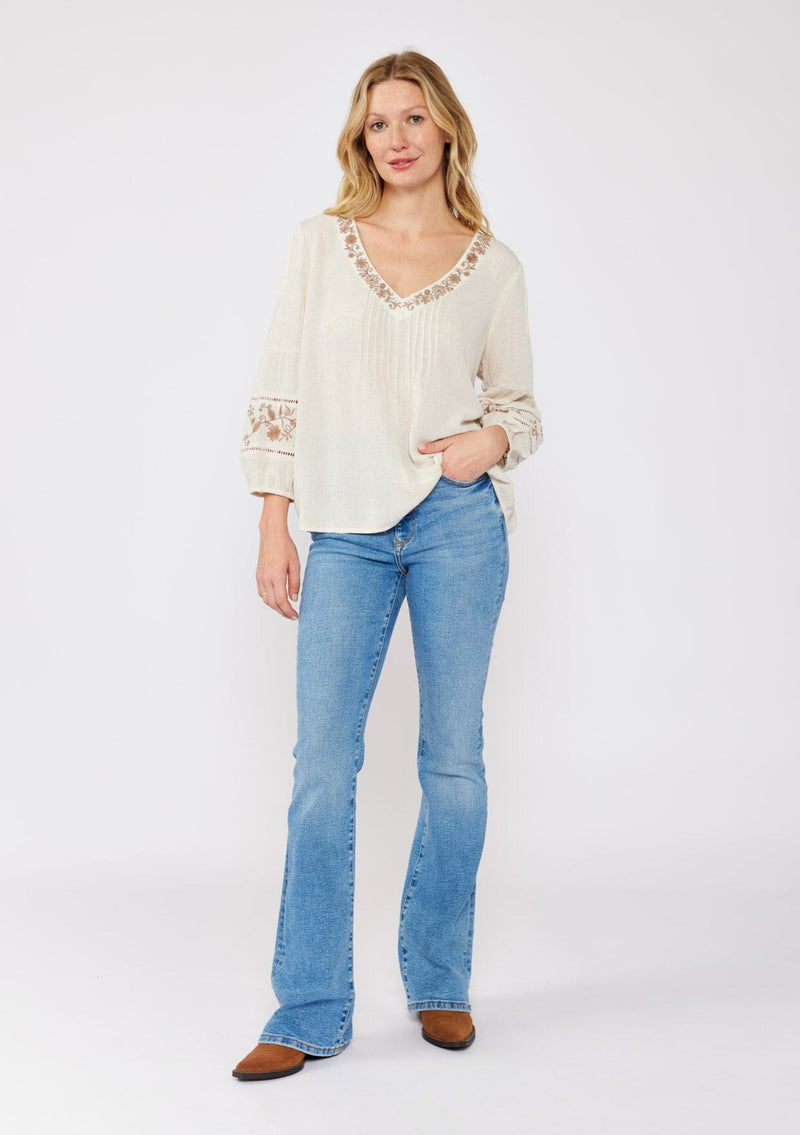 [Color: Natural/Brown] An image of a blonde model wearing an ultra bohemian blouse with embroidered details. A beige natural tone blouse with a v neckline, floral embroidery, and long sleeves with elastic cuffs. A relaxed fit fall season top paired with denim jeans.