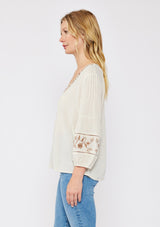 [Color: Natural/Brown] An image of a blonde model wearing an ultra bohemian blouse with embroidered details. A beige natural tone blouse with a v neckline, floral embroidery, and long sleeves with elastic cuffs. A relaxed fit fall season top paired with denim jeans.