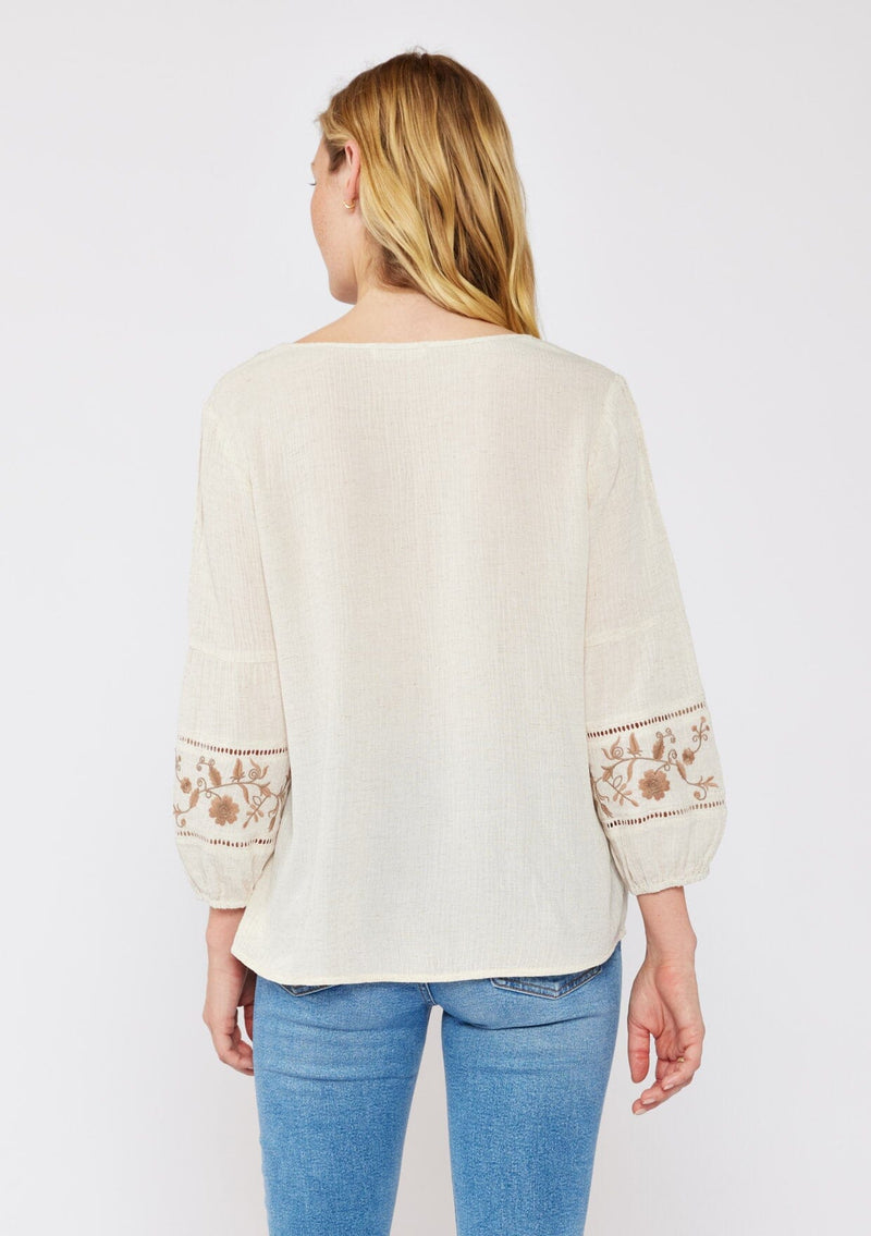 [Color: Natural/Brown] An image of a blonde model wearing an ultra bohemian blouse with embroidered details. A beige natural tone blouse with a v neckline, floral embroidery, and long sleeves with elastic cuffs. A relaxed fit fall season top paired with denim jeans.