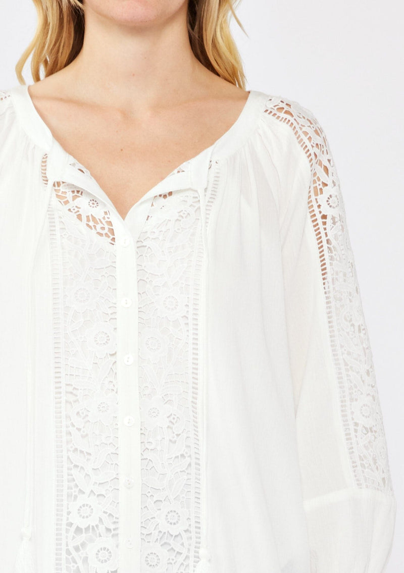 [Color: Cream] A blonde model wearing an off white bohemian resort blouse with voluminous long sleeves, a button front, tassel neck ties, and lace trim throughout.
