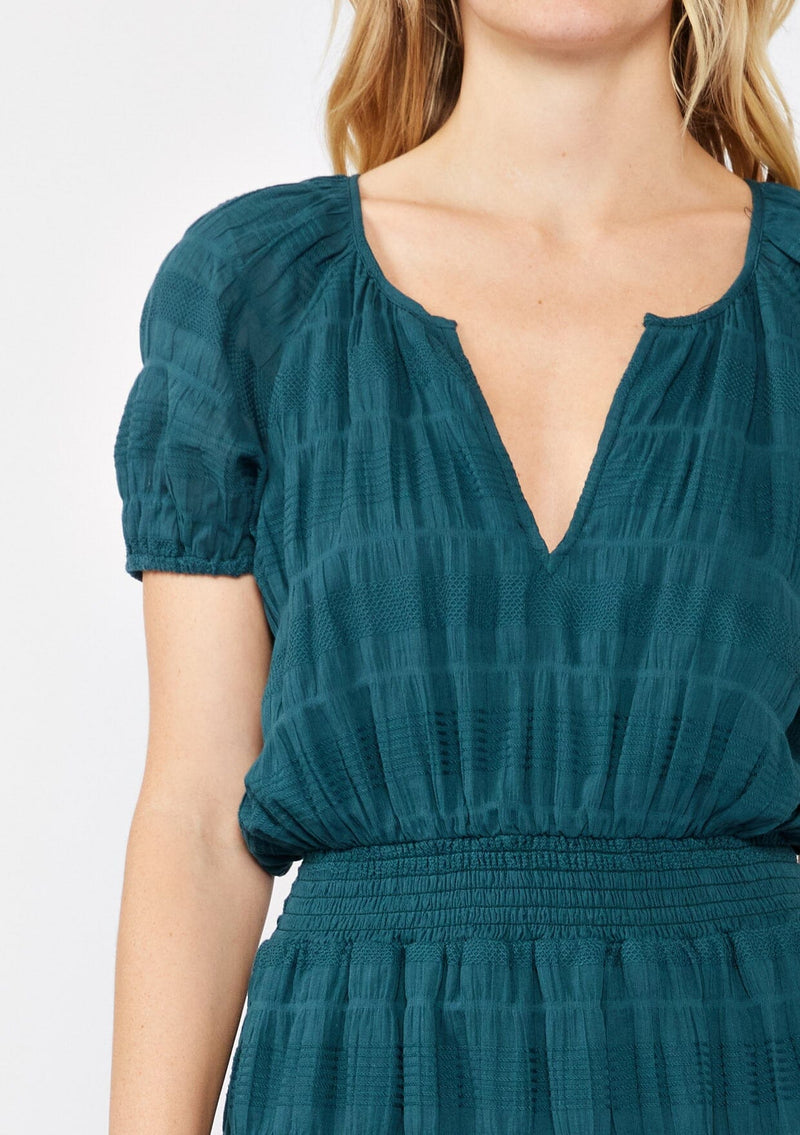 [Color: Deep Teal] A blonde model wearing a teal midi dress with a subtle woven striped design. With a split v-neckline, short puff sleeves, smocked waistline, and a ruffle trimmed hem.