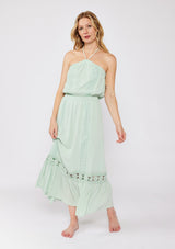 [Color: Dusty Sage] A blonde model wearing a minty green ultra bohemian maxi dress for vacation. This summer maxi dress features a halter neckline and a comfortable smocked waist. This dress is also embellished with intricate lace details and embroidered eyelet. The perfect style for brunch or a tropical vacation.