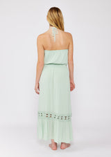 [Color: Dusty Sage] A blonde model wearing a minty green ultra bohemian maxi dress for vacation. This summer maxi dress features a halter neckline and a comfortable smocked waist. This dress is also embellished with intricate lace details and embroidered eyelet. The perfect style for brunch or a tropical vacation.