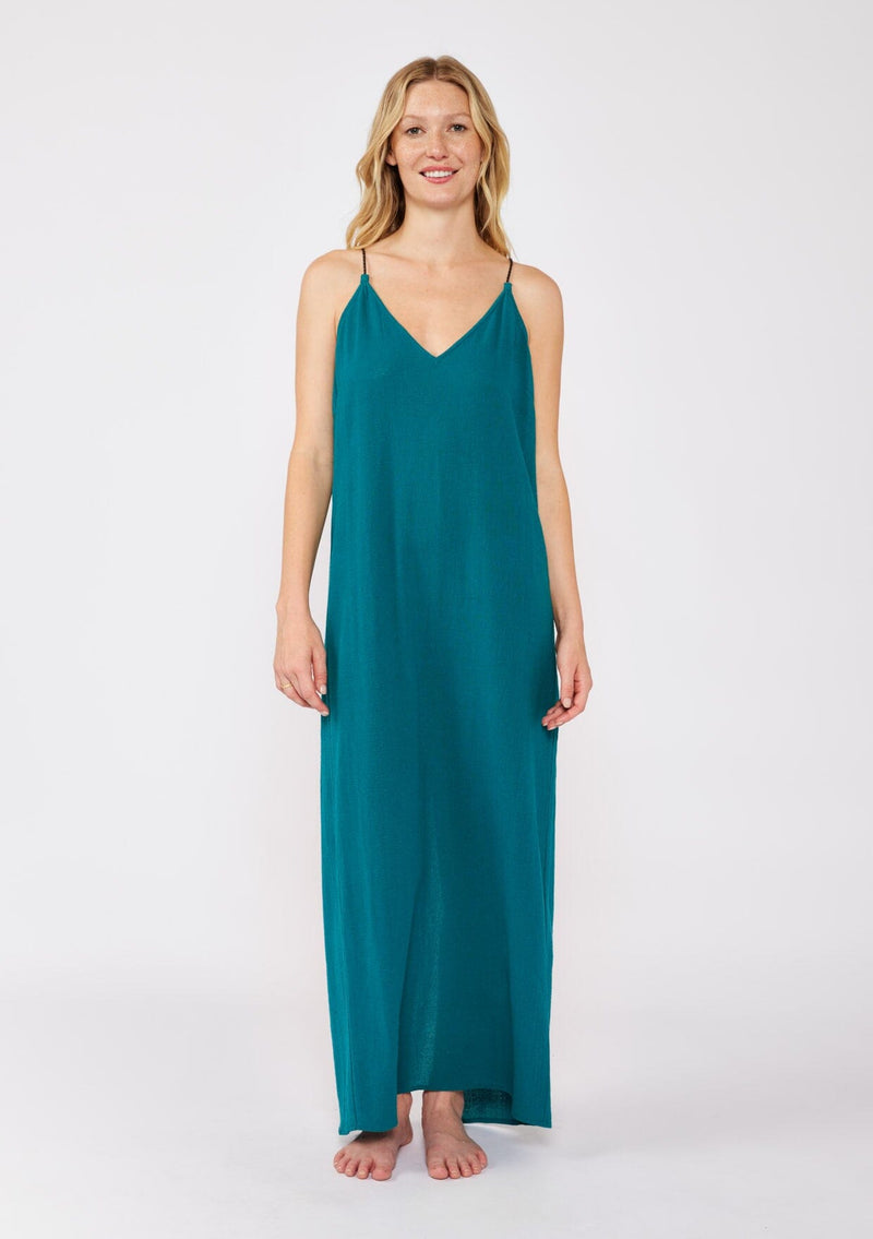 [Color: Forest Teal] A blonde model wearing a flowy, casual summer dress in a teal solid print. With a v neckline, relaxed fit, and faux braided leather straps that are adjustable at the back. A cute back slit dress for the summer season, perfect for vacation or beachside stroll. Classic cotton dress.