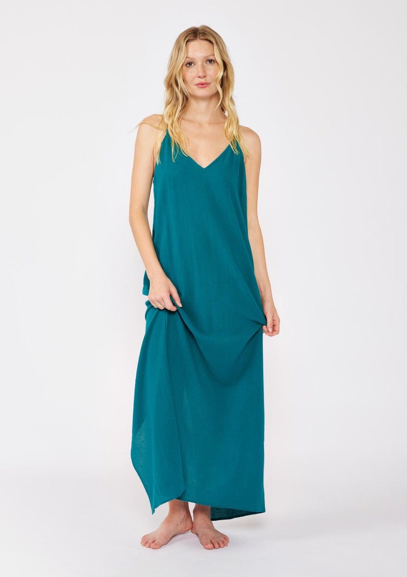 [Color: Forest Teal] A blonde model wearing a flowy, casual summer dress in a teal solid print. With a v neckline, relaxed fit, and faux braided leather straps that are adjustable at the back. A cute back slit dress for the summer season, perfect for vacation or beachside stroll. Classic cotton dress.