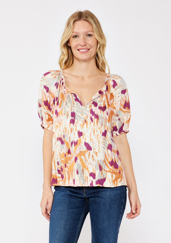 [Color: Natural/Plum] A blonde model wearing an orange and purple abstract ikat print blouse. With a split v neckline with ties, short puff sleeves, and lace trim details at the sleeve. A fall bohemian top styled with dark denim jeans. 