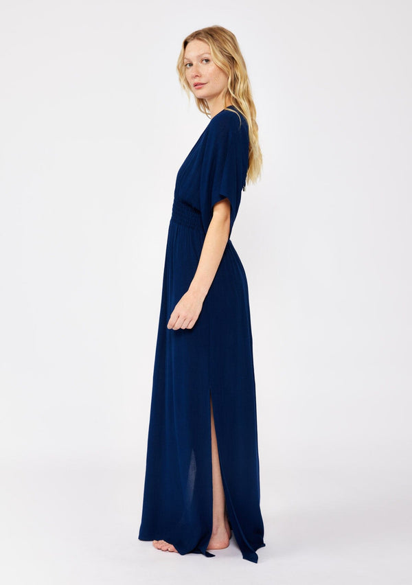 [Color: Pacific Blue] A side facing image of a blonde model wearing a resort ready blue navy maxi dress. With half length kimono sleeves, a plunging v neckline, a smocked elastic empire waist, side slits, and an open back with tie closure.