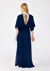 [Color: Pacific Blue] A back facing image of a blonde model wearing a resort ready blue navy maxi dress. With half length kimono sleeves, a plunging v neckline, a smocked elastic empire waist, side slits, and an open back with tie closure.