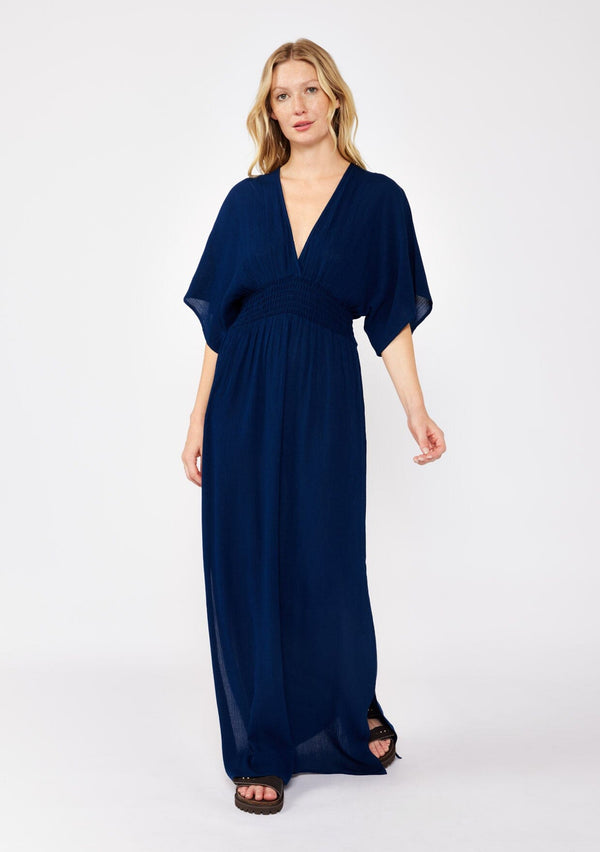 [Color: Pacific Blue] A front facing image of a blonde model wearing a resort ready blue navy maxi dress. With half length kimono sleeves, a plunging v neckline, a smocked elastic empire waist, side slits, and an open back with tie closure.