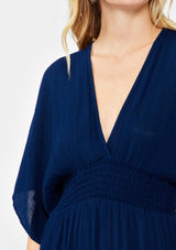 [Color: Pacific Blue] A detailed image of a blonde model wearing a resort ready blue navy maxi dress. With half length kimono sleeves, a plunging v neckline, a smocked elastic empire waist, side slits, and an open back with tie closure.