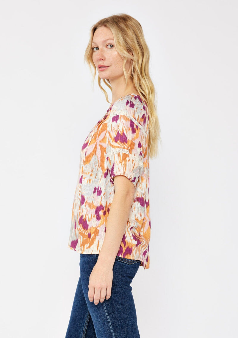 [Color: Natural/Plum] A blonde model wearing an orange and purple abstract ikat print blouse. With a split v neckline with ties, short puff sleeves, and lace trim details at the sleeve. A fall bohemian top styled with dark denim jeans.