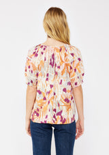 [Color: Natural/Plum] A blonde model wearing an orange and purple abstract ikat print blouse. With a split v neckline with ties, short puff sleeves, and lace trim details at the sleeve. A fall bohemian top styled with dark denim jeans.