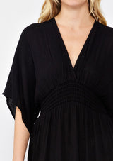 [Color: Black] A front facing image of a blonde model wearing a resort ready black maxi dress. With half length kimono sleeves, a plunging v neckline, a smocked elastic empire waist, side slits, and an open back with tie closure.