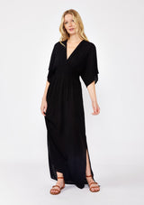 [Color: Black] A front facing image of a blonde model wearing a resort ready black maxi dress. With half length kimono sleeves, a plunging v neckline, a smocked elastic empire waist, side slits, and an open back with tie closure.