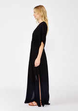 [Color: Black] A front facing image of a blonde model wearing a resort ready black maxi dress. With half length kimono sleeves, a plunging v neckline, a smocked elastic empire waist, side slits, and an open back with tie closure.