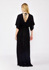 [Color: Black] A front facing image of a blonde model wearing a resort ready black maxi dress. With half length kimono sleeves, a plunging v neckline, a smocked elastic empire waist, side slits, and an open back with tie closure.