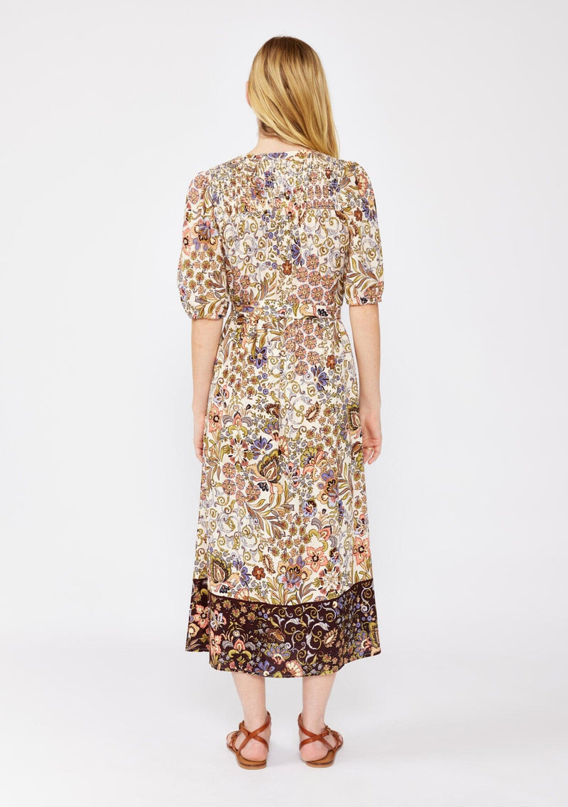 [Color: Natural/Brown] A blonde model wearing an ultra-bohemian fall midi dress in a brown floral border print. With a functional button front, adjustable waist belt, and puff sleeves with elastic cuffs. An autumn day dress styled with strappy flats for a casual fall look.