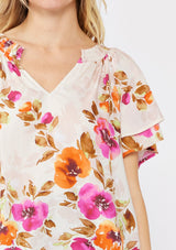 [Color: Natural/Orchid] A blonde model wearing a relaxed fit top in a floral print. With short double flutter sleeves, split v neckline, and a smocked detail at the neck. A dressy but casual floral top styled with dark denim.