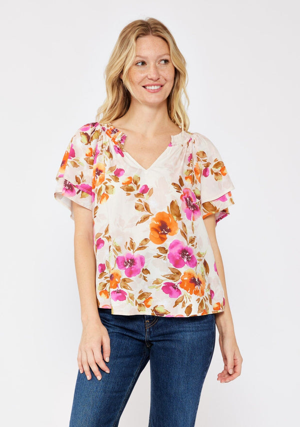 [Color: Natural/Orchid] A blonde model wearing a relaxed fit top in a floral print. With short double flutter sleeves, split v neckline, and a smocked detail at the neck. A dressy but casual floral top styled with dark denim. 