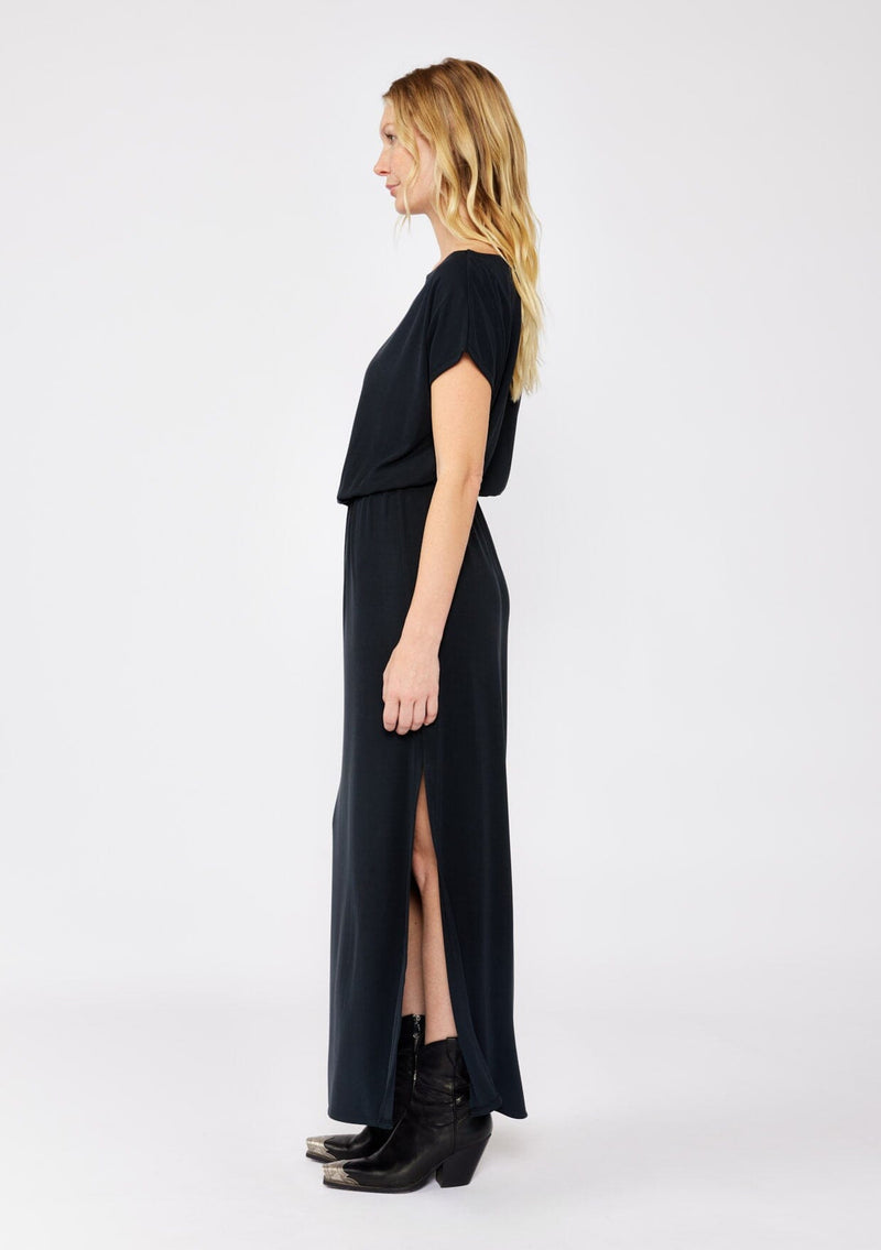 [Color: Black] A blonde model wearing a relaxed fit maxi that can be worn on or off shoulder. A casual black dress with a boat neckline, short dolman sleeves, elastic waistline, and side slits. Styled with black western boots for the fall season.