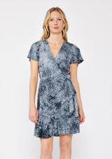 [Color: Indigo] A blonde model wearing an abstract animal print mini wrap dress in a blue hue. With a side tie closure, flutter short sleeves, and a ruffle trimmed hem. A fall wrap dress styled with black boots.