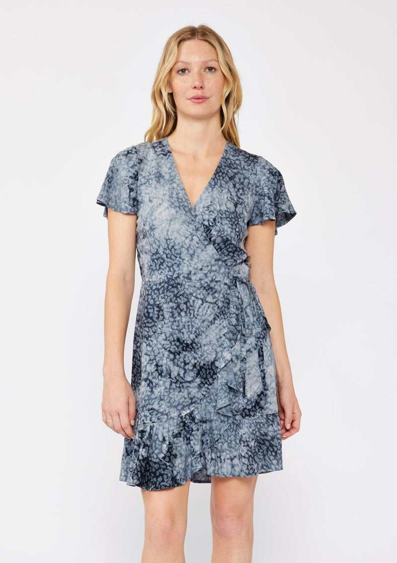 [Color: Indigo] A blonde model wearing an abstract animal print mini wrap dress in a blue hue. With a side tie closure, flutter short sleeves, and a ruffle trimmed hem. A fall wrap dress styled with black boots.