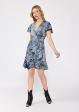 [Color: Indigo] A blonde model wearing an abstract animal print mini wrap dress in  a blue hue. With a side tie closure, flutter short sleeves, and a ruffle trimmed hem. A fall wrap dress styled with black boots. 