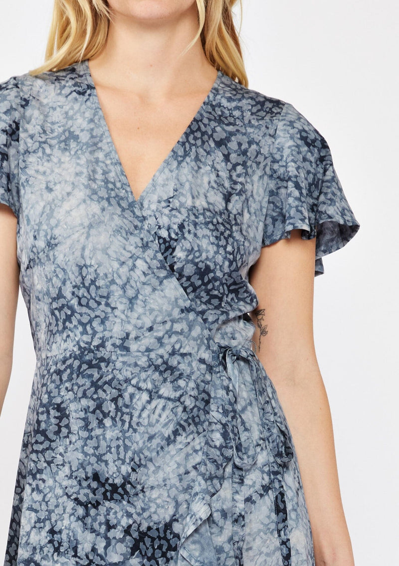 [Color: Indigo] A blonde model wearing an abstract animal print mini wrap dress in a blue hue. With a side tie closure, flutter short sleeves, and a ruffle trimmed hem. A detailed image of a fall wrap dress.