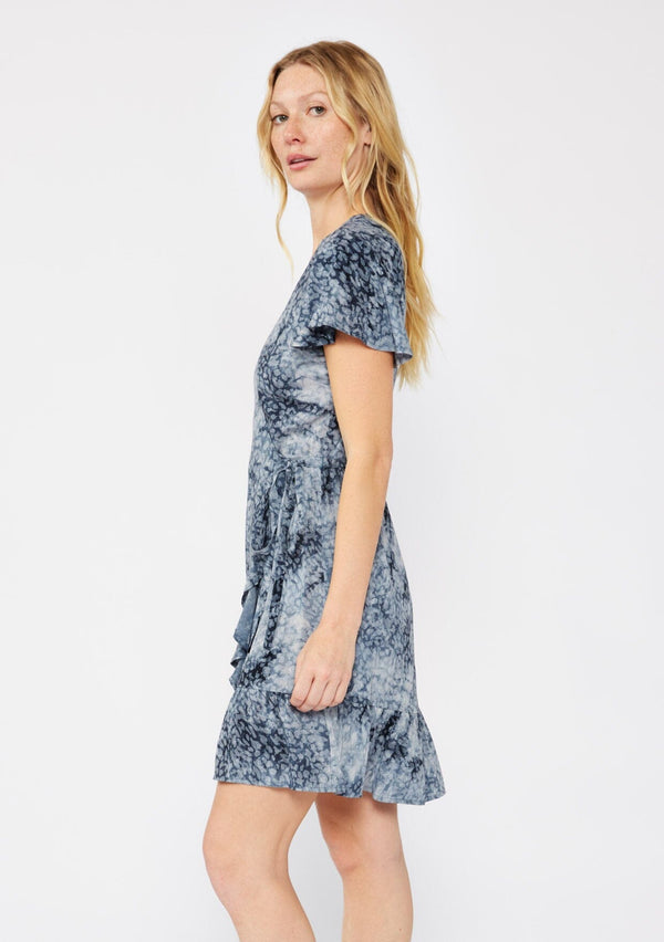 [Color: Indigo] A blonde model wearing an abstract animal print mini wrap dress in a blue hue. With a side tie closure, flutter short sleeves, and a ruffle trimmed hem. A fall wrap dress styled with black boots.