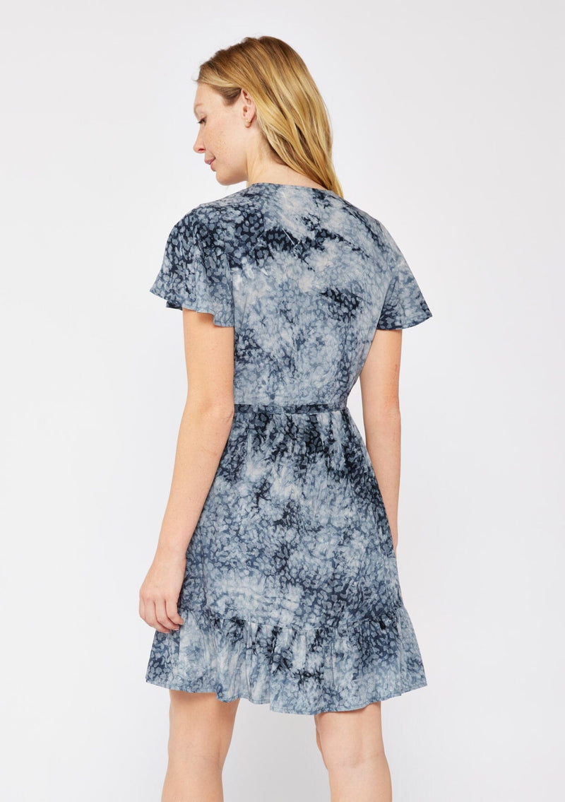 [Color: Indigo] A blonde model wearing an abstract animal print mini wrap dress in a blue hue. With a side tie closure, flutter short sleeves, and a ruffle trimmed hem. A fall wrap dress styled with black boots.