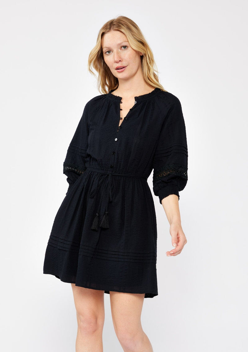 [Color: Black] A blonde model wearing a ultra fall bohemian mini dress in black. With a button front, long sleeves with smocked cuffs, tassel tie waist detail, and lace inserts.